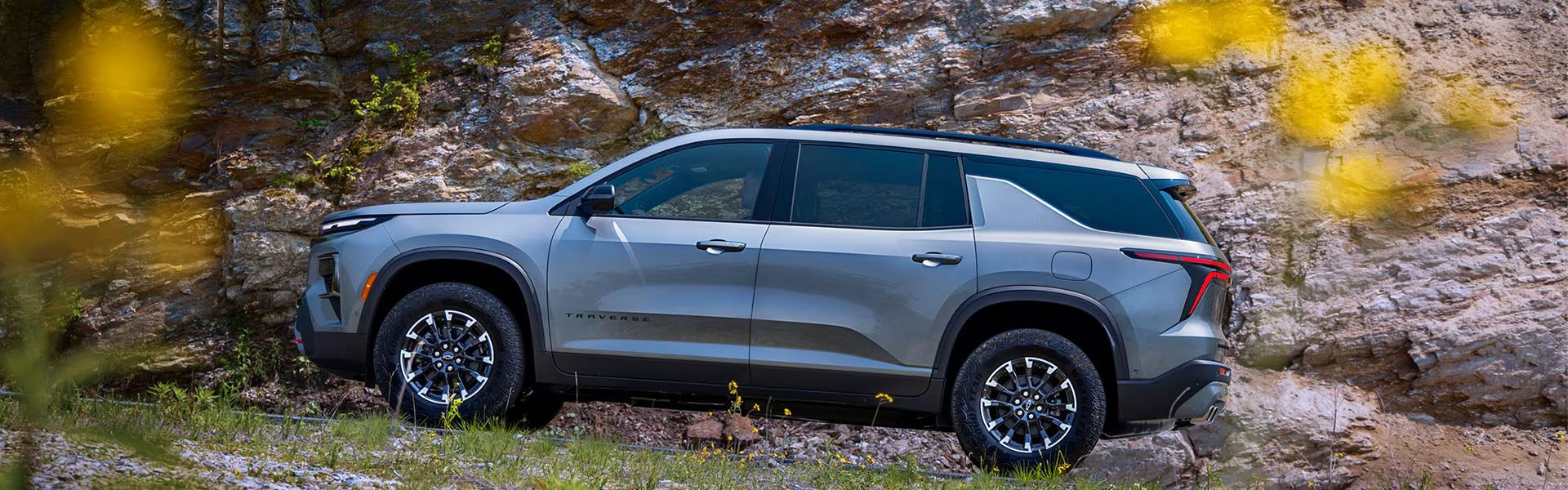 Gray 2026 Chevrolet Traverse parked on a rugged roadside near rocky terrain, shown in side profile with black wheels and surrounding greenery.