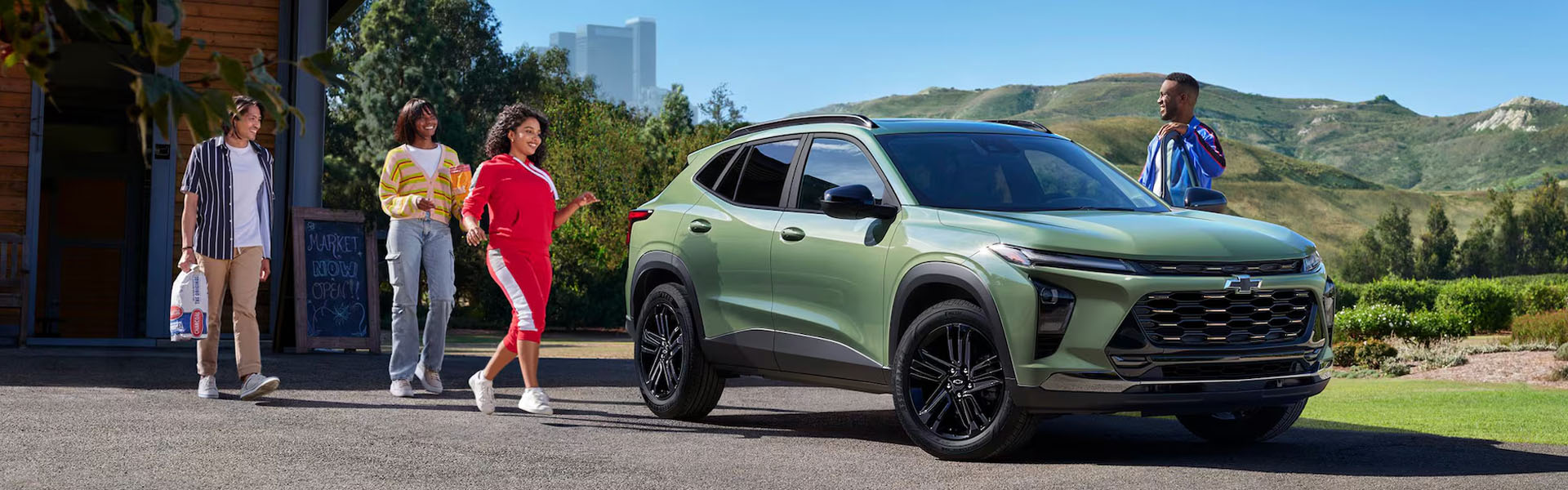 Green 2026 Chevrolet Trax parked near a small market, with a group of people walking nearby and scenic hills in the background under a clear sky.