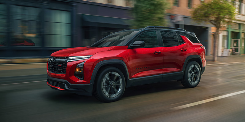 A sleek, red Equinox drives down a wet city street. The vehicle's modern design and shiny finish create a sense of speed and contemporary style.
